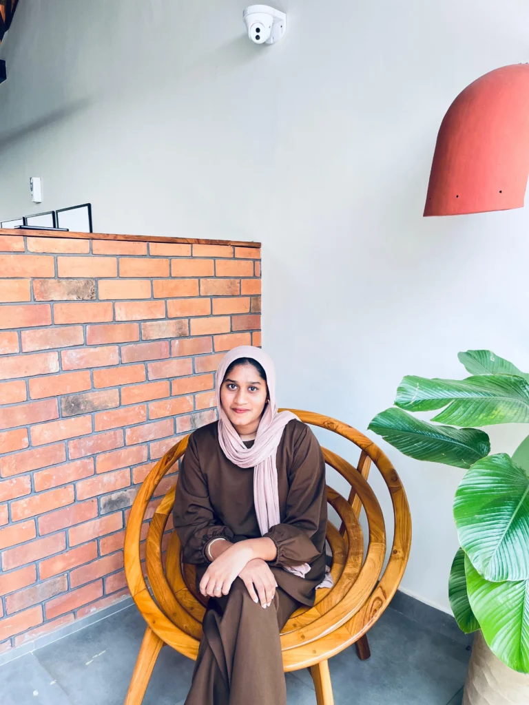 A digital marketer in a brown outfit with a light pink hijab sitting on a wooden chair near a brick wall and green plant indoors. Best Freelance Digital Marketing Analyst in calicut